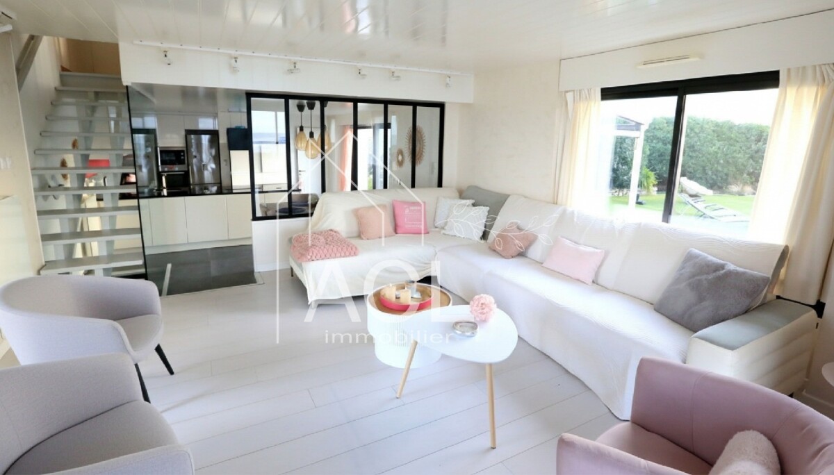 Bright open-plan living room with a white L-shaped sofa, pastel pink cushions, and a pale lavender accent chair; large glass doors to a green garden outside.