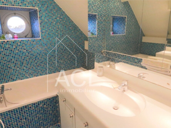 Blue mosaic-tiled bathroom with a white bathtub along the wall and a double sink vanity under a large mirror. A round window and skylight illuminate the room.