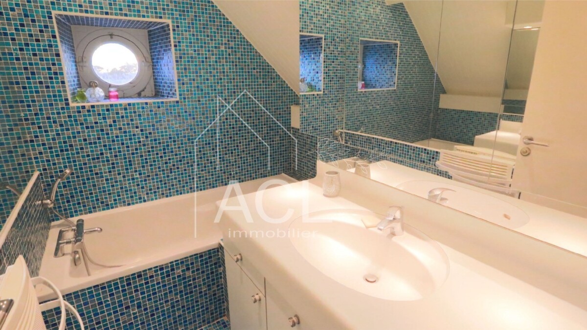 Blue mosaic-tiled bathroom with a white bathtub along the wall and a double sink vanity under a large mirror. A round window and skylight illuminate the room.