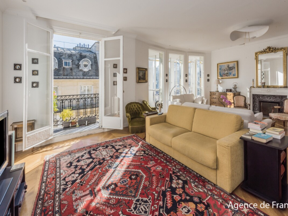 Bright living room with a beige sofa, red oriental rug, and French doors opening to a balcony overlooking city rooftops.