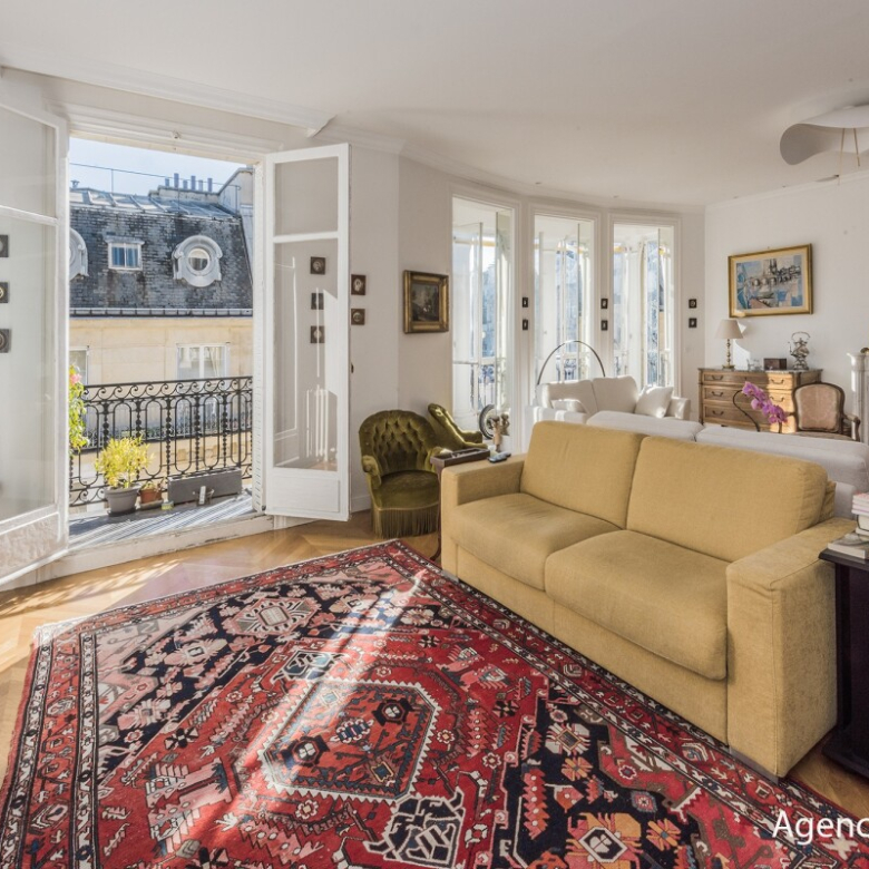 Bright living room with a beige sofa, red oriental rug, and French doors opening to a balcony overlooking city rooftops.