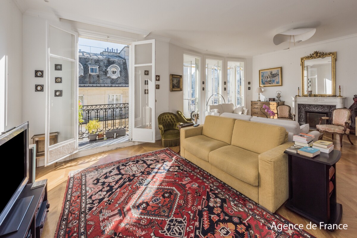 Bright living room with a beige sofa, red oriental rug, and French doors opening to a balcony overlooking city rooftops.