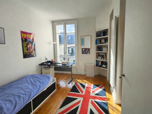 Bright bedroom with a single bed on the left, a black desk by a large window, and a Union Jack rug on a wooden floor, with a bookcase corner.