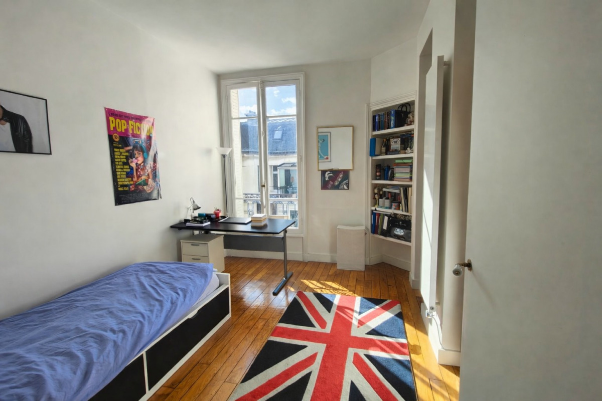 Bright bedroom with a single bed on the left, a black desk by a large window, and a Union Jack rug on a wooden floor, with a bookcase corner.