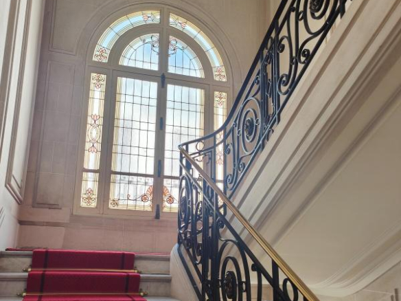 Marble staircase with a red carpet runner and decorative black wrought-iron railing, ascending toward a large arched stained-glass window.