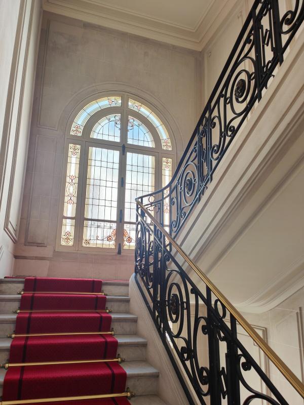 Marble staircase with a red carpet runner and decorative black wrought-iron railing, ascending toward a large arched stained-glass window.