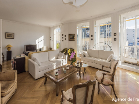 Bright living room with two white sofas, vintage armchairs, and a wooden coffee table, opening to large windows and a city view.