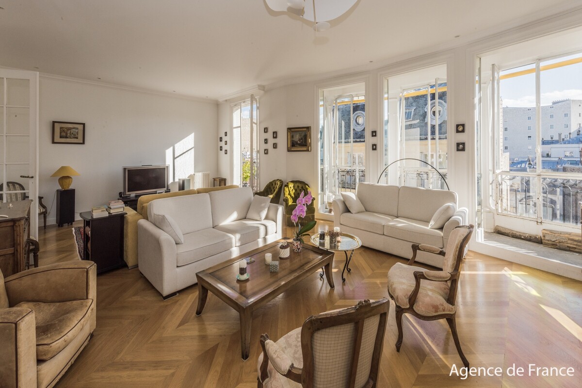Bright living room with two white sofas, vintage armchairs, and a wooden coffee table, opening to large windows and a city view.