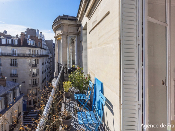 Parisian balcony with bright blue folding chairs and potted plants overlooking a city street under a clear sky.