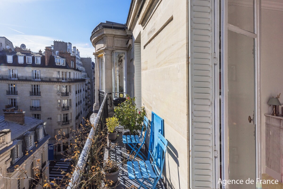 Parisian balcony with bright blue folding chairs and potted plants overlooking a city street under a clear sky.