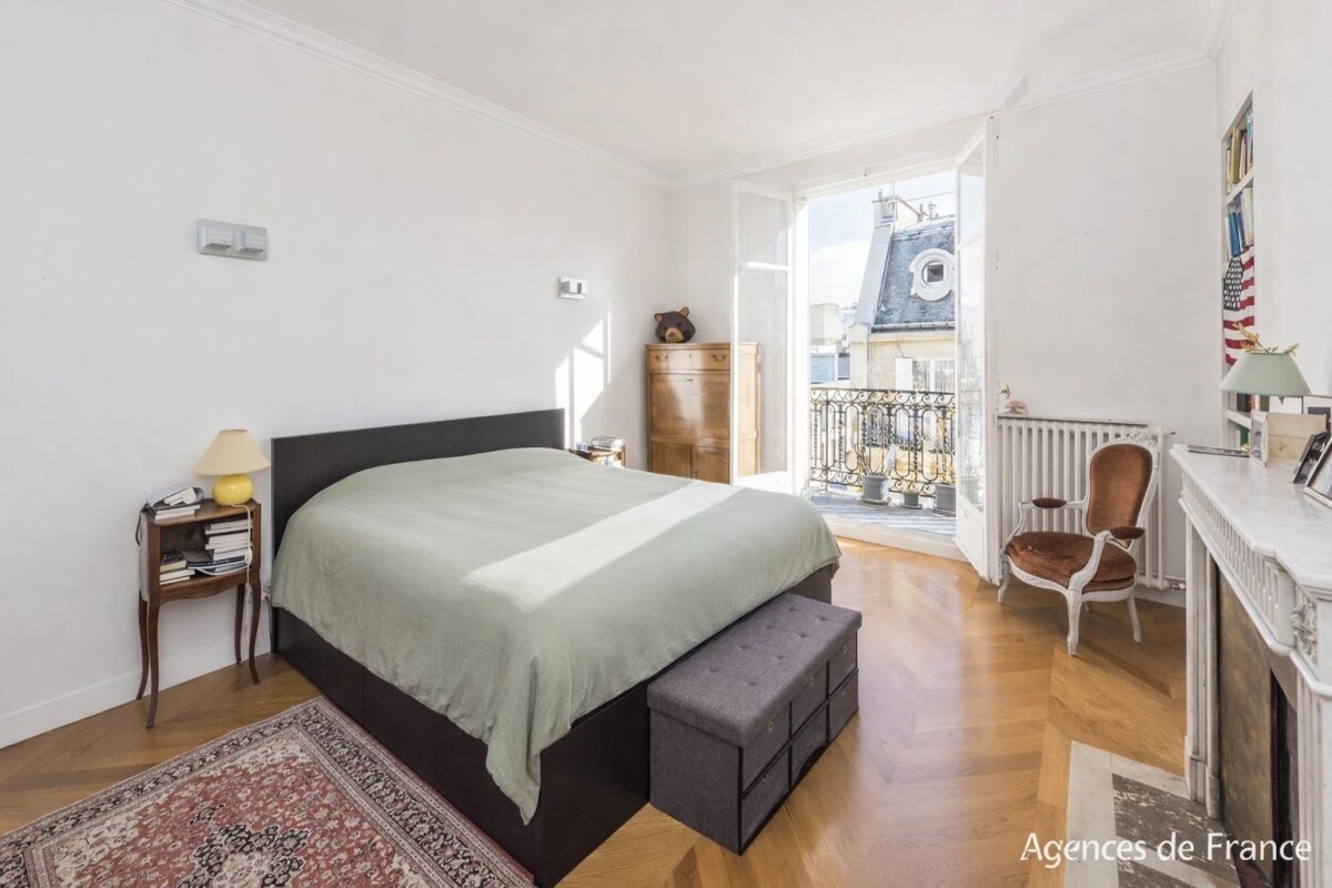 Bright bedroom with an open French balcony door, a large bed, and a green bedspread; a bench, nightstand with books, and a wooden dresser on the left.
