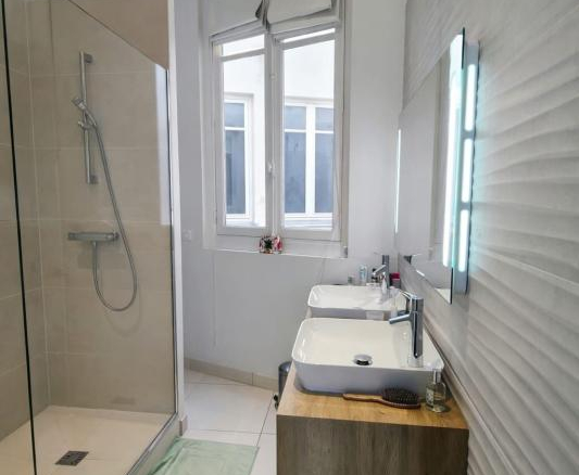 Modern bathroom with a glass shower enclosure, a wood vanity with two white vessel sinks, a large window, and mint-green bath mats.