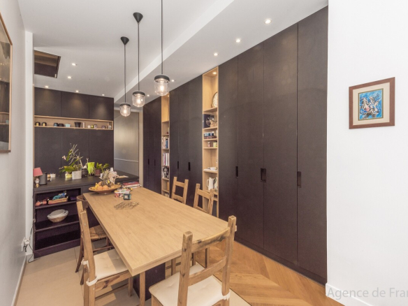 Modern dining room with a long wooden table, six wooden chairs, and tall dark storage cabinets along the wall, illuminated by pendant lights.