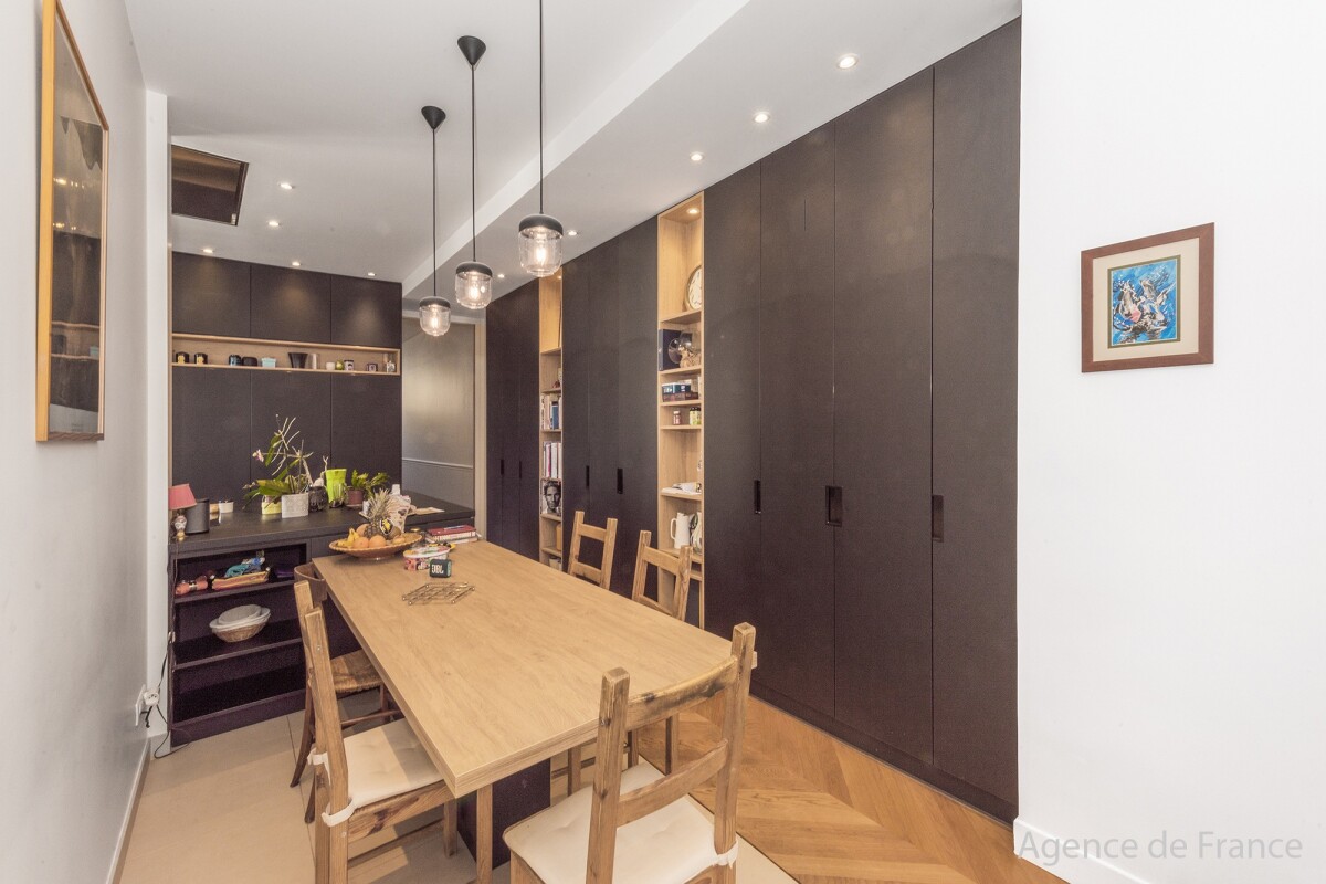 Modern dining room with a long wooden table, six wooden chairs, and tall dark storage cabinets along the wall, illuminated by pendant lights.