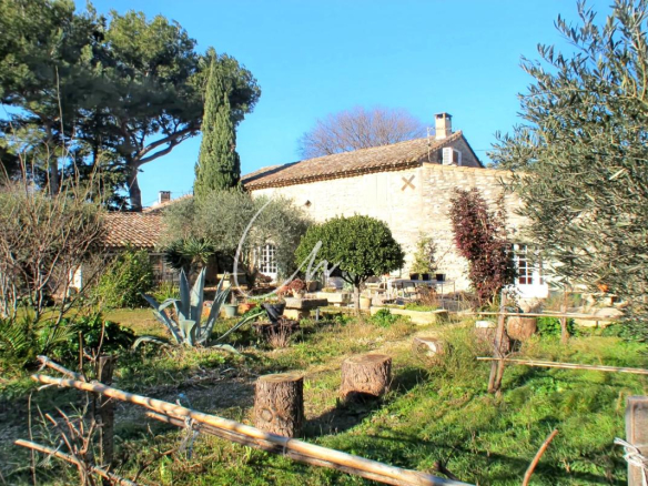 Stone cottage with tiled roof in a sunny garden, featuring outdoor seating and potted plants in front.