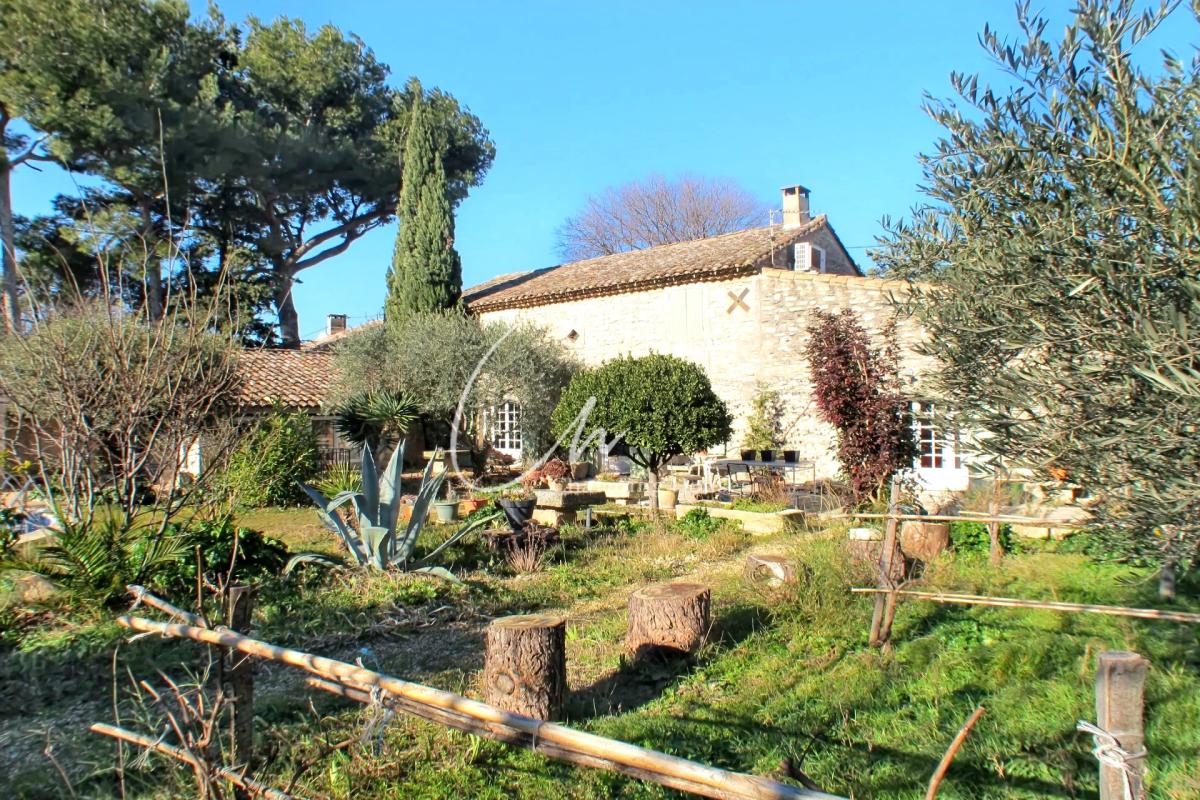 Stone cottage with tiled roof in a sunny garden, featuring outdoor seating and potted plants in front.