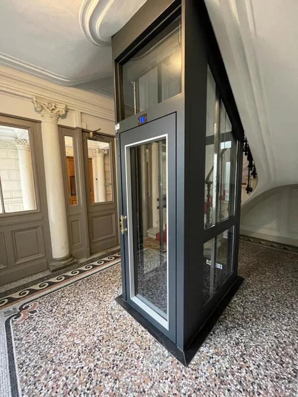 Ascenseur vitré dans un hall d'immeuble bourgeois à Paris, avec des murs en pierre et un sol en mosaïque.