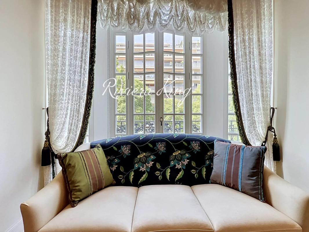 Intérieur d'un appartement Belle Époque avec un canapé, des rideaux élégants et des fenêtres ouvertes sur l'extérieur.