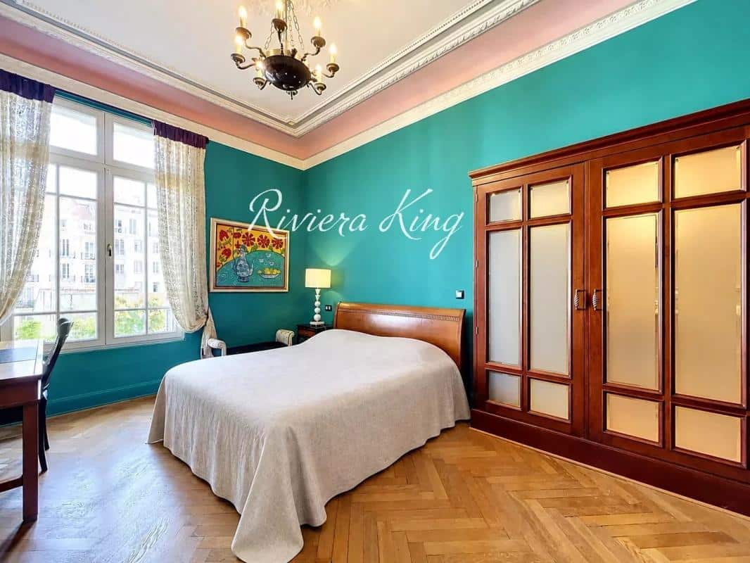 Chambre d'un appartement Belle Époque avec murs turquoise et parquet, fenêtres lumineuses, mobilier en bois.