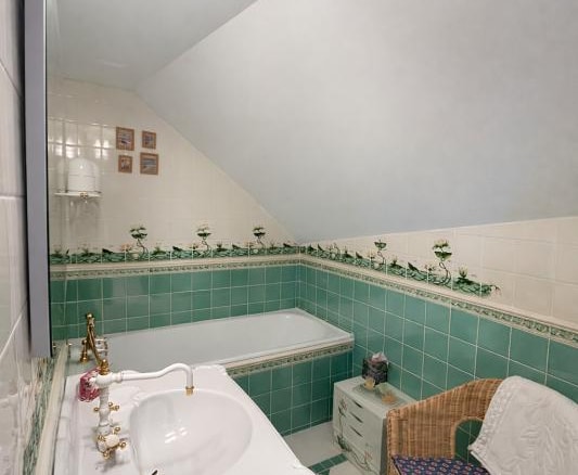 Salle de bain d'un triplex avec baignoire, lavabo et chaise en osier, décorée de carrelage vert.