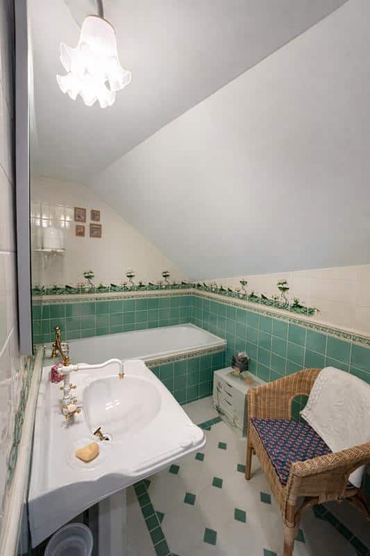 Salle de bain d'un triplex avec baignoire, lavabo et chaise en osier, décorée de carrelage vert.