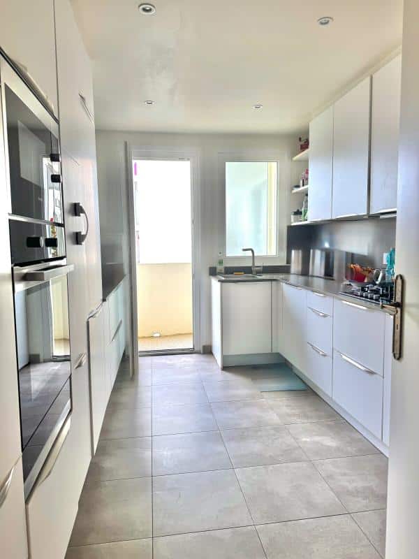 Modern kitchen in a Biarritz apartment with white furniture and windows opening onto a balcony.