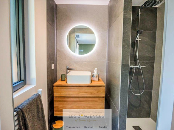 Modern bathroom with shower, washbasin on wooden unit and illuminated round mirror.