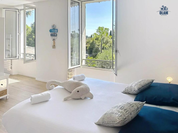 Chambre lumineuse avec fenêtres ouvertes, décorée de coussins bleus et d'une peluche en forme de poisson.
