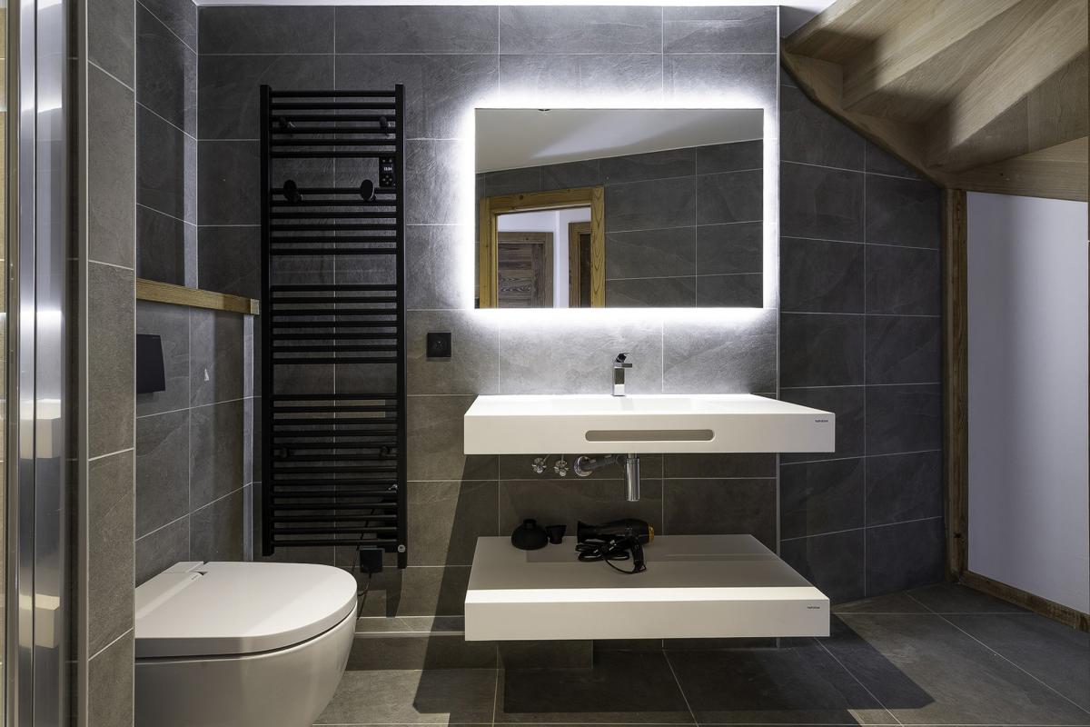 Salle de bain moderne avec lavabo suspendu et douche, carrelage gris, miroir lumineux.