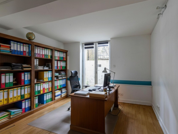 Office with a bookcase and a wooden desk, windows overlooking the outside.
