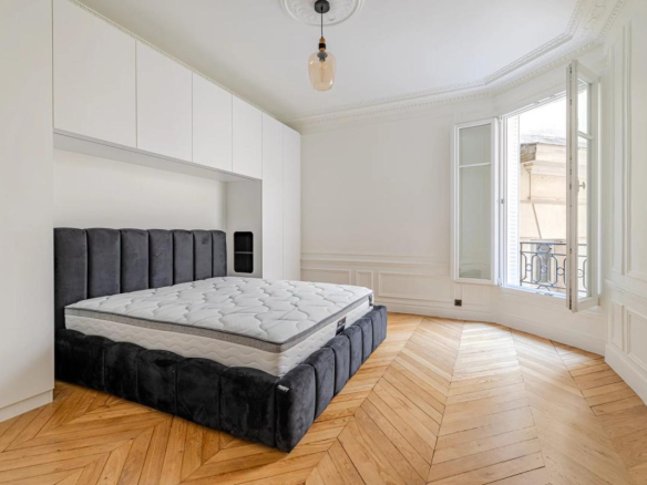 Room in a renovated apartment with herringbone parquet flooring and an unmade bed, windows open to the outside.