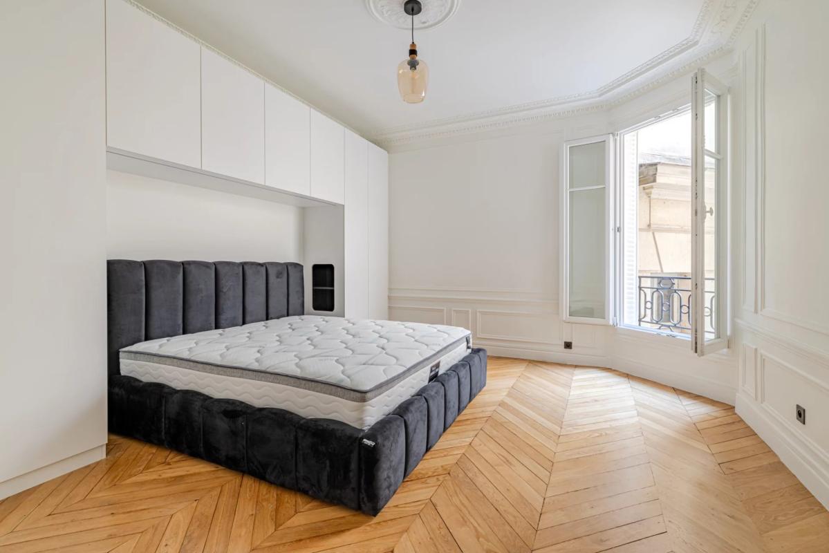 Room in a renovated apartment with herringbone parquet flooring and an unmade bed, windows open to the outside.
