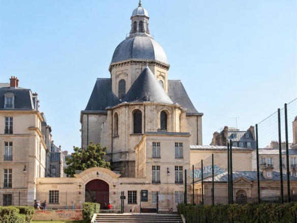 Église avec dôme et croix, vue depuis un espace extérieur avec des arbres et un terrain de sport.