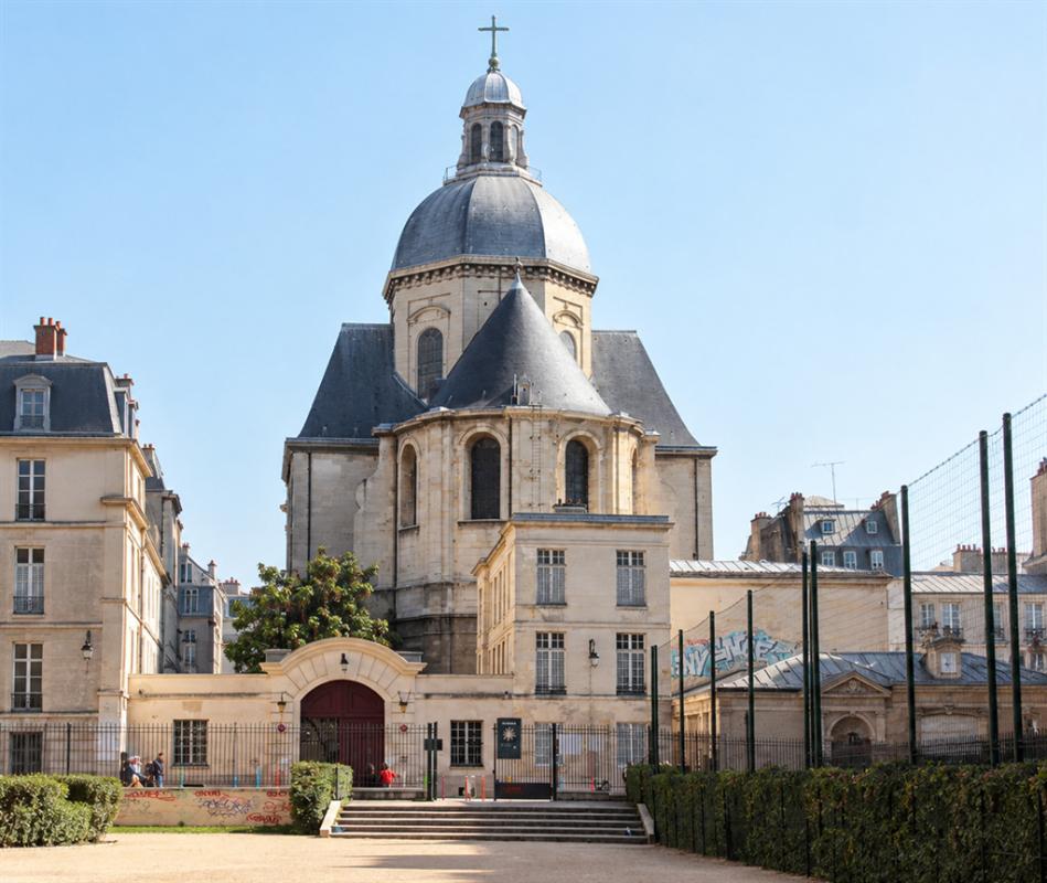 Église dans le Marais à Paris Église avec dôme et croix, vue depuis un espace extérieur avec des arbres et un terrain de sport.