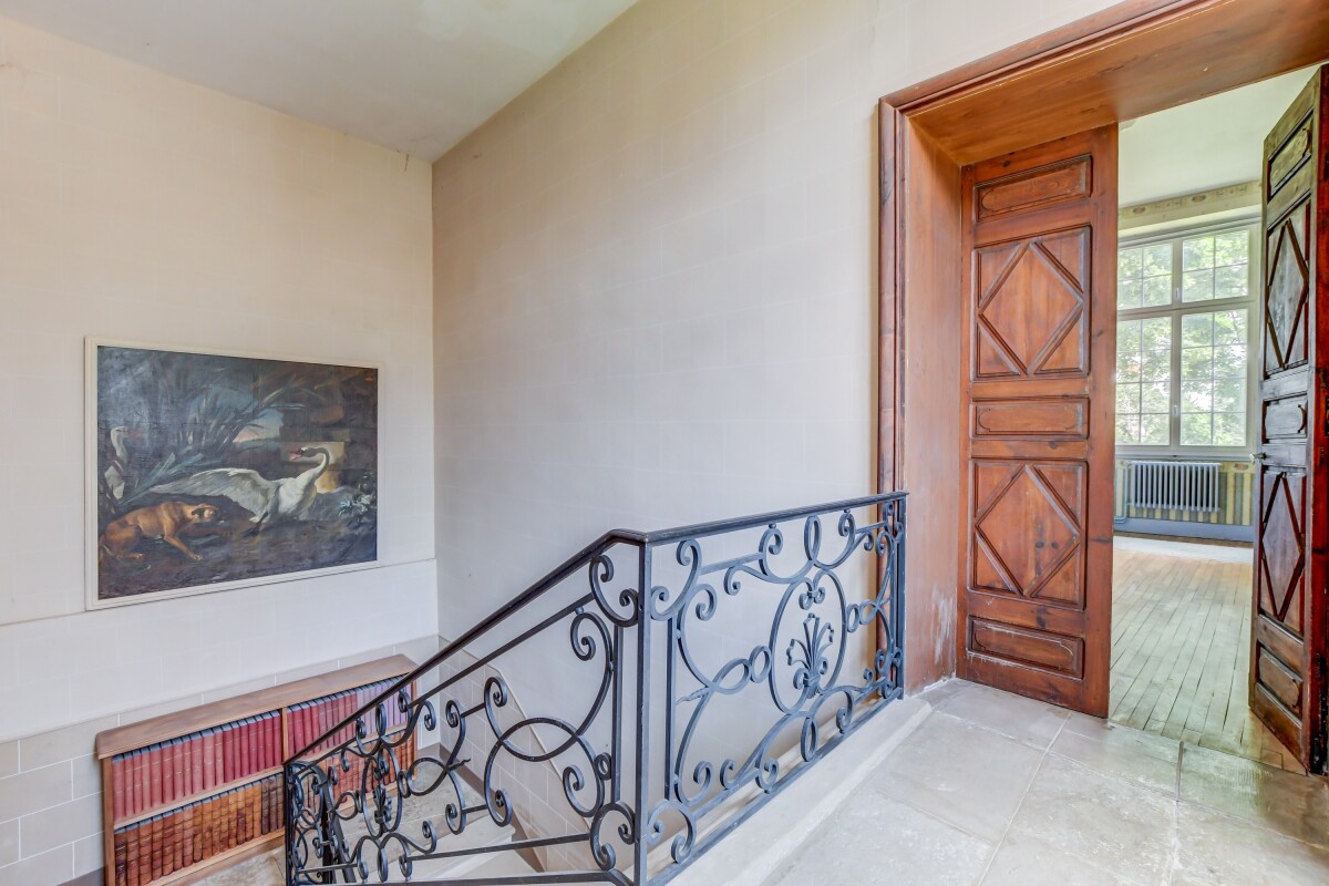 Intérieur d’un château à Lanta Escalier avec balustrade en fer forgé menant à un espace lumineux, avec une peinture accrochée au mur.
