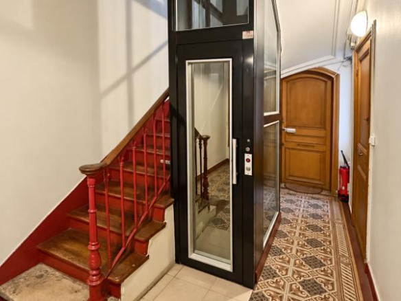 Glass elevator in a Haussmannian building lobby with staircase and decorative tiling.