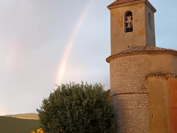 Église avec clocher et arbre, sous un ciel partiellement nuageux avec un arc-en-ciel à Coursegoules.
