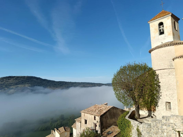 Vue panoramique sur un village avec une église et des montagnes en arrière-plan, sous un ciel dégagé.