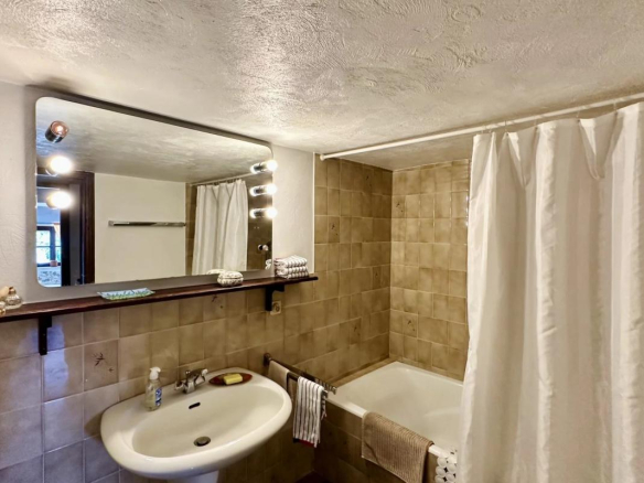 Salle de bain avec baignoire, lavabo et miroir, murs carrelés en beige.