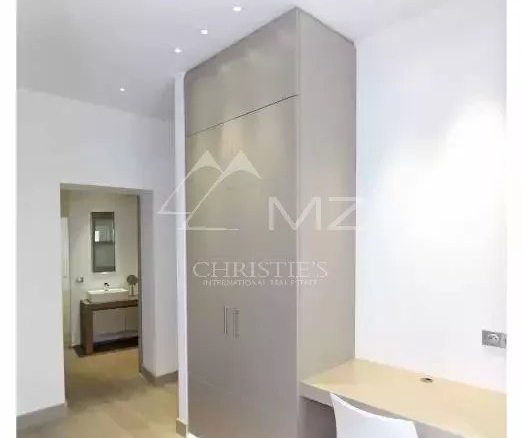 Modern room with a tall gray built-in cabinet, a light wooden desk, and a white chair; doorway to a bathroom visible in the background.
