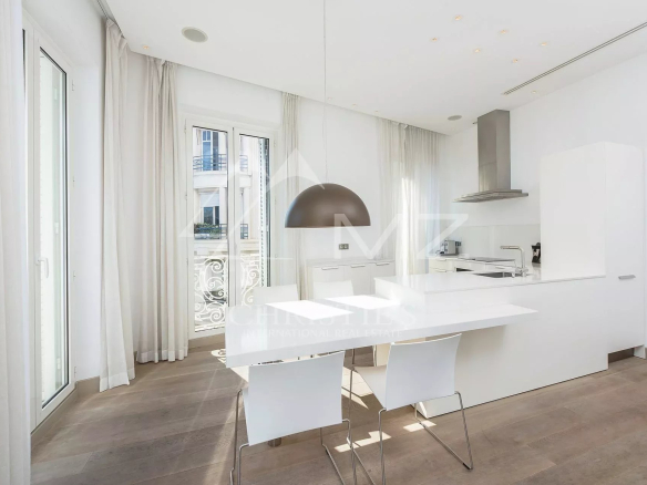 Bright white minimalist kitchen with a long white island, white chairs, and a large arched pendant light over the dining area near glass doors to a balcony.
