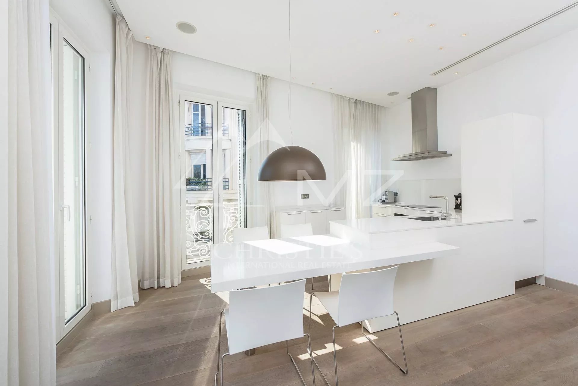 Bright white minimalist kitchen with a long white island, white chairs, and a large arched pendant light over the dining area near glass doors to a balcony.