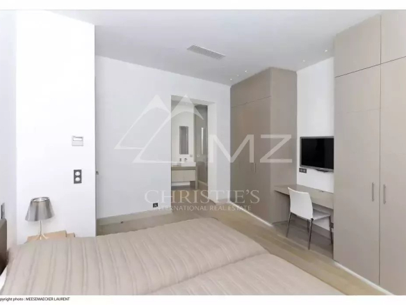 Modern bedroom with a large wall of beige built-in wardrobes, a wall-mounted TV above a small desk and white chair, and a neatly made beige bed in the foreground.
