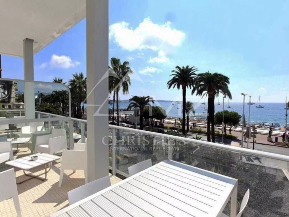 Outdoor seating area with white tables and chairs on a terrace overlooking a palm-lined promenade and the blue sea.
