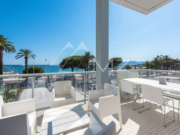 Seaside balcony with white dining table and chairs, glass railing, and a view of palm trees and the blue sea.