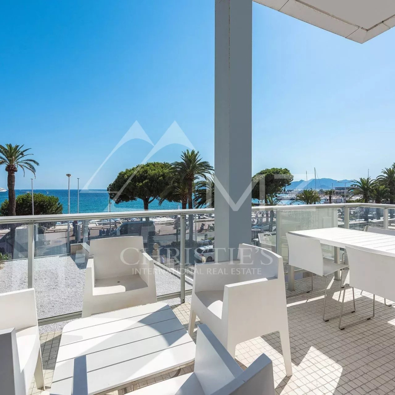 Seaside balcony with white dining table and chairs, glass railing, and a view of palm trees and the blue sea.