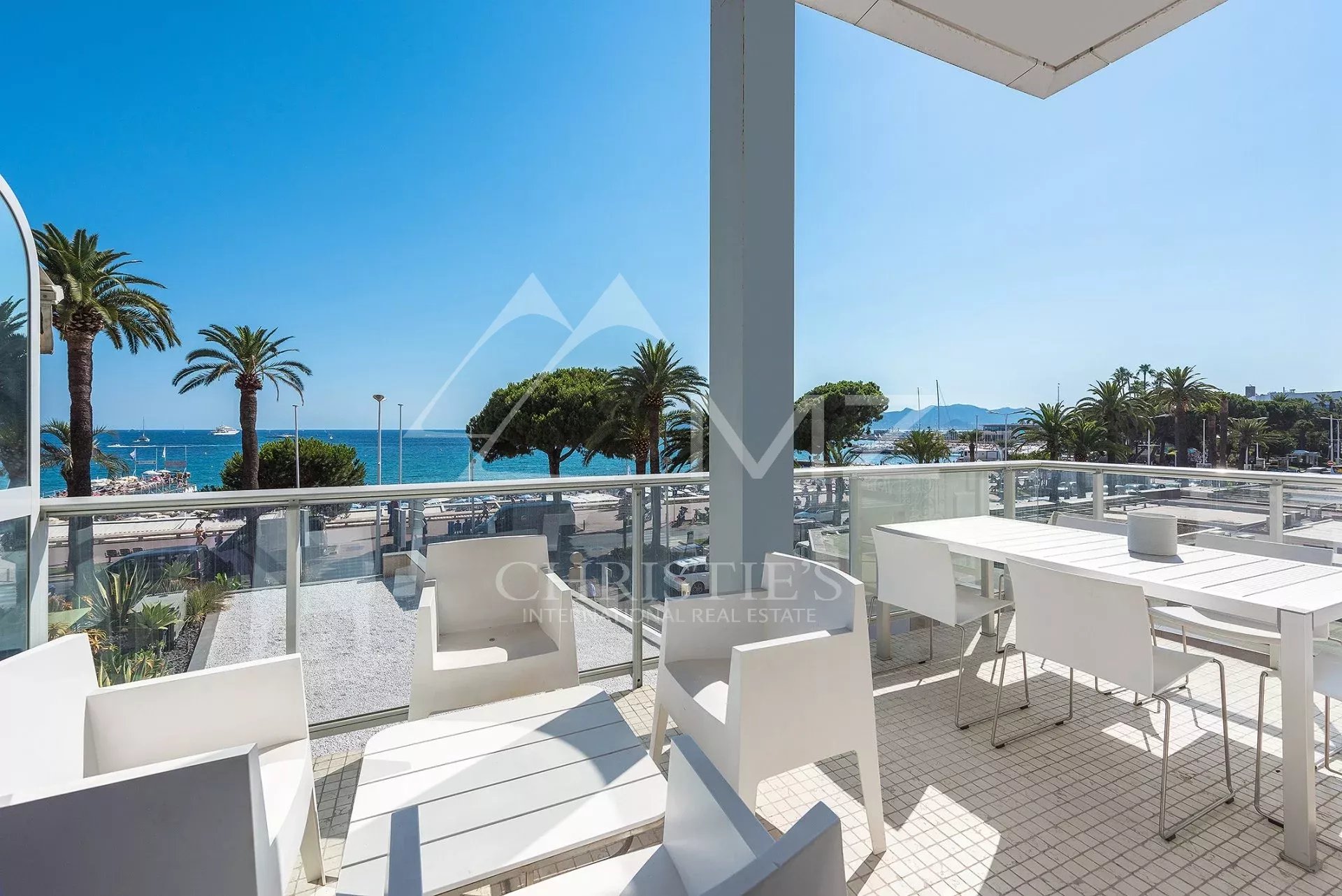 Seaside balcony with white dining table and chairs, glass railing, and a view of palm trees and the blue sea.