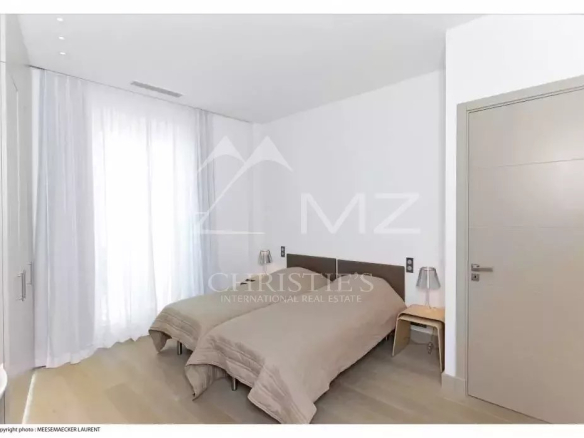 Bright modern bedroom with two single beds, beige duvets, nightstands, and a large window with sheer white curtains.