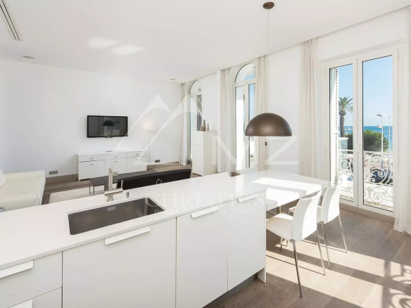 Bright open-plan kitchen and living area in a white, modern apartment, with a large island sink, white cabinetry, and a dining table near glass doors leading to a balcony with a sea view