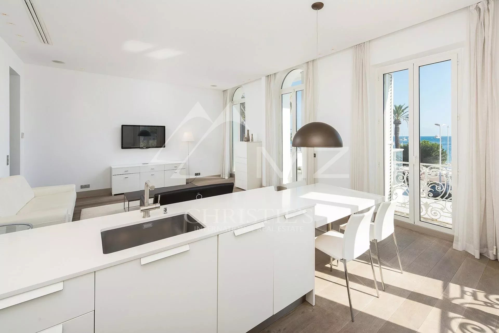 Bright open-plan kitchen and living area in a white, modern apartment, with a large island sink, white cabinetry, and a dining table near glass doors leading to a balcony with a sea view